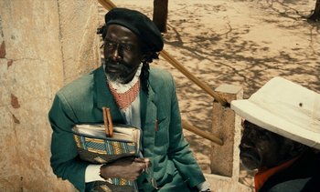 Movie still from “Hyenas” (1992), directed by Djibril Diop Mambéty – A man sitting on steps holding a book in his hands; Medium shot, Over the shoulder angle