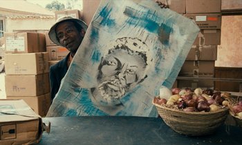 Movie still from “Hyenas” (1992), directed by Djibril Diop Mambéty – A man holding up a painting of a face; Close Up shot, Over the shoulder angle