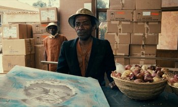 Movie still from “Hyenas” (1992), directed by Djibril Diop Mambéty – A man sitting in front of a table with a basket of onions; Medium shot, Over the shoulder angle