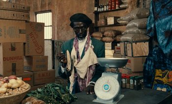 Movie still from “Hyenas” (1992), directed by Djibril Diop Mambéty – A man standing at a table with a scale; Medium shot, High angle