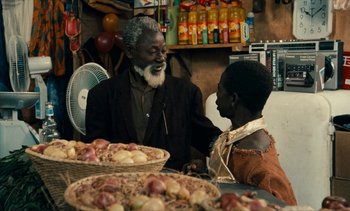 Movie still from “Hyenas” (1992), directed by Djibril Diop Mambéty – A man sitting next to a young boy in front of baskets of fruit; Medium shot, Over the shoulder angle