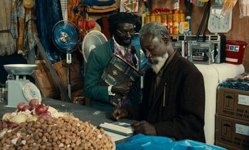 Movie still from “Hyenas” (1992), directed by Djibril Diop Mambéty – Two men are working together in a market; Medium shot, Over the shoulder angle