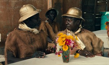 Movie still from “Hyenas” (1992), directed by Djibril Diop Mambéty – A group of men sitting at a table with a vase of flowers; Medium shot, Over the shoulder angle