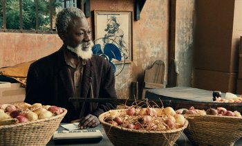 Movie still from “Hyenas” (1992), directed by Djibril Diop Mambéty – An older man sitting at a table with a basket of fruit; Medium shot, Over the shoulder angle