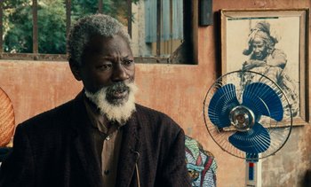 Movie still from “Hyenas” (1992), directed by Djibril Diop Mambéty – An older man sitting in front of an electric fan; Close Up shot, Over the shoulder angle