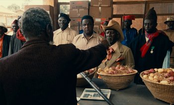 Movie still from “Hyenas” (1992), directed by Djibril Diop Mambéty – A group of men standing in front of a basket of food; Medium shot, Over the shoulder angle