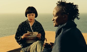 Movie still from “Hyenas” (1992), directed by Djibril Diop Mambéty – Two people sitting on the beach looking at each other; Medium shot, Over the shoulder angle
