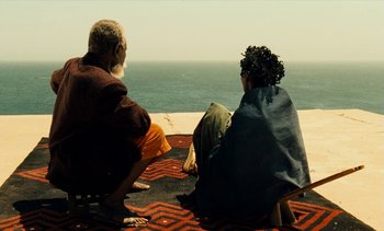 Movie still from “Hyenas” (1992), directed by Djibril Diop Mambéty – Two people sitting on the ground looking out at the ocean; Wide shot, High angle