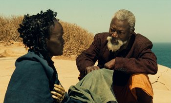 Movie still from “Hyenas” (1992), directed by Djibril Diop Mambéty – A man and a woman sitting on the ground talking to each other; Close Up shot, Over the shoulder angle