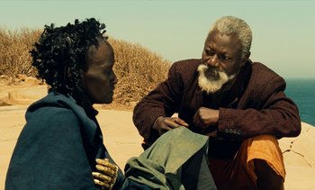 Movie still from “Hyenas” (1992), directed by Djibril Diop Mambéty – An older man and a younger woman in a desert setting; Close Up shot, Over the shoulder angle
