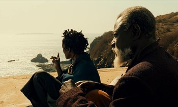 Movie still from “Hyenas” (1992), directed by Djibril Diop Mambéty – A man and a woman sitting on the beach; Medium shot, High angle