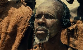 Movie still from “Hyenas” (1992), directed by Djibril Diop Mambéty – An older man with a beard and headphones on; Close Up shot, Low angle