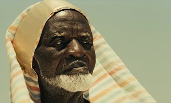 Movie still from “Hyenas” (1992), directed by Djibril Diop Mambéty – An old man with a beard and a head covering; Close Up shot, Low angle