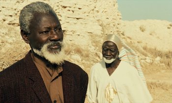 Movie still from “Hyenas” (1992), directed by Djibril Diop Mambéty – Two older men standing next to each other in front of a cliff; Close Up shot, Over the shoulder angle