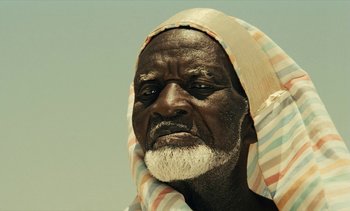 Movie still from “Hyenas” (1992), directed by Djibril Diop Mambéty – An old man with a white beard wearing a towel around his neck; Close Up shot, Low angle