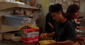 Movie still from “Jason's Lyric” (1994), directed by Doug McHenry – A woman in black shirt cutting a box of chips; Medium shot, Over the shoulder angle