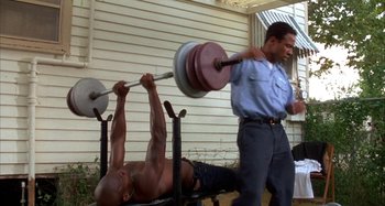 Movie still from “Jason's Lyric” (1994), directed by Doug McHenry – A man lifting a barbell over a bench with another man standing next to it; Medium shot, Low angle