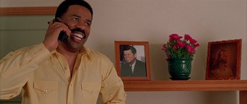 Movie still from “Johnson Family Vacation” (2004), directed by Christopher Erskin – A man smiling in front of a picture of john f; Medium shot, Low angle