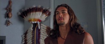 Movie still from “Johnson Family Vacation” (2004), directed by Christopher Erskin – A man with long brown hair and a long beard standing in front of an indian headdress; Close Up shot, Over the shoulder angle