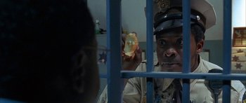 Movie still from “Johnson Family Vacation” (2004), directed by Christopher Erskin – A police officer is holding a cookie in a jail cell; Close Up shot, Over the shoulder angle