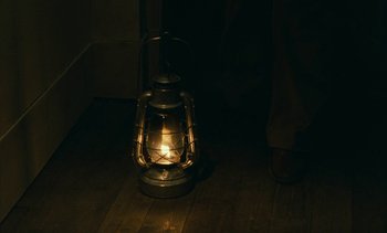 Movie still from “L'Argent” (1983), directed by Robert Bresson – An old lantern is lit on the floor of a dark room; Wide shot, High angle