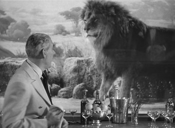 Movie still from “Mighty Joe Young” (1949), directed by Ernest B. Schoedsack – A man sitting at a table with a lion in the background; Medium shot, Over the shoulder angle
