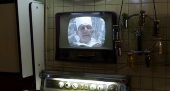 Movie still from “Mood Indigo” (2013), directed by Michel Gondry – A television with a man on it in the middle of a kitchen; Close Up shot, Over the shoulder angle