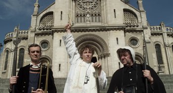 Movie still from “Mood Indigo” (2013), directed by Michel Gondry – A man in a white robe holds up a trumpet in front of a building; Medium shot, Low angle