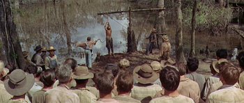 Movie still from “Nevada Smith” (1966), directed by Henry Hathaway – A group of people in the woods watching a man swing a stick; Extreme Wide shot, High angle