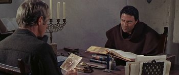 Movie still from “Nevada Smith” (1966), directed by Henry Hathaway – A man sitting at a table reading a book to another man; Medium shot, Over the shoulder angle