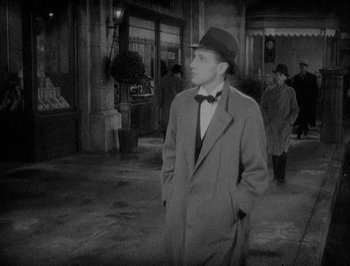 Movie still from “Of Human Bondage” (1934), directed by John Cromwell – A man wearing a hat and a bow tie standing on a sidewalk; Medium shot, High angle