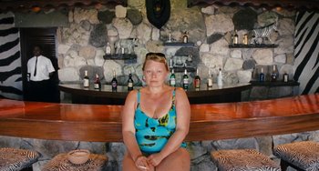 Movie still from “Paradise: Love” (2012), directed by Ulrich Seidl – A woman sitting in front of a bar with bottles on the wall; Medium shot, High angle