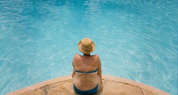 Movie still from “Paradise: Love” (2012), directed by Ulrich Seidl – A woman in a straw hat sitting on the edge of a swimming pool; Wide shot, Overhead angle