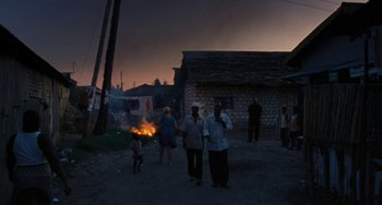 Movie still from “Paradise: Love” (2012), directed by Ulrich Seidl – A group of people standing on a dirt road near a fire; Extreme Wide shot, High angle