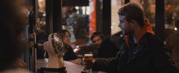 Movie still from “Simon Killer” (2012), directed by Antonio Campos – A man sitting at a table holding a glass of beer in front of two other people; Medium shot, Over the shoulder angle