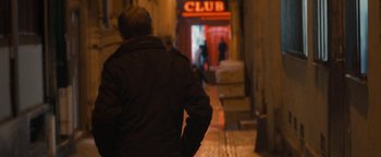 Movie still from “Simon Killer” (2012), directed by Antonio Campos – A man walking down a street at night past a club sign; Medium shot, Over the shoulder angle