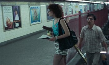 Movie still from “Smithereens” (1982), directed by Susan Seidelman – A woman walking down a hallway holding papers and an apple; Wide shot, Over the shoulder angle