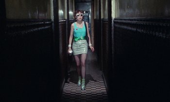 Movie still from “Smithereens” (1982), directed by Susan Seidelman – A woman walking down a hall way wearing sunglasses and holding a drink; Wide shot, Low angle