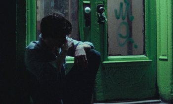 Movie still from “Smithereens” (1982), directed by Susan Seidelman – A man sitting on the ground in front of a green door; Close Up shot, High angle