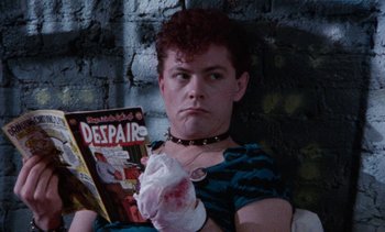 Movie still from “Smithereens” (1982), directed by Susan Seidelman – A man sitting in front of a brick wall holding a comic book; Close Up shot, High angle