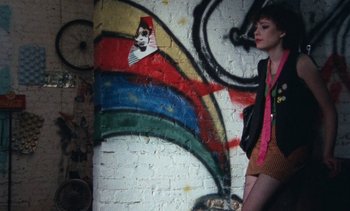 Movie still from “Smithereens” (1982), directed by Susan Seidelman – A woman standing in front of a wall with a painting on it; Wide shot, Overhead angle