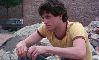 Movie still from “Smithereens” (1982), directed by Susan Seidelman – A man sitting on top of a rock near a brick wall; Close Up shot, Low angle
