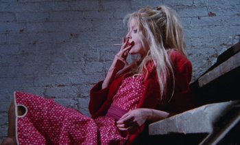 Movie still from “Smithereens” (1982), directed by Susan Seidelman – A woman sitting down smoking a cigarette in front of a brick wall; Medium shot, Low angle