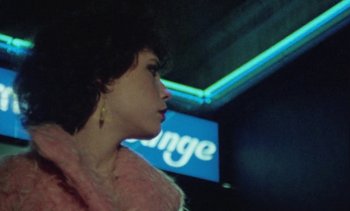 Movie still from “Smithereens” (1982), directed by Susan Seidelman – A person in a room with a neon sign in the background; Close Up shot, Over the shoulder angle