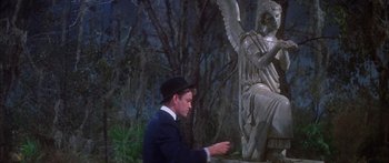 Movie still from “Summer and Smoke” (1961), directed by Peter Glenville – A man wearing a suit and a hat standing in front of a statue; Medium shot, Over the shoulder angle