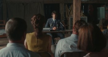 Movie still from “Taking Woodstock” (2009), directed by Ang Lee – A man sitting at a table in front of a microphone; Medium shot, Over the shoulder angle