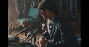 Movie still from “Taking Woodstock” (2009), directed by Ang Lee – A man sitting at a table with microphones in front of him; Medium shot, Over the shoulder angle