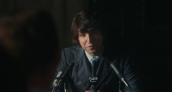 Movie still from “Taking Woodstock” (2009), directed by Ang Lee – A man wearing a suit and tie while sitting at a microphone; Close Up shot, Over the shoulder angle