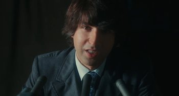 Movie still from “Taking Woodstock” (2009), directed by Ang Lee – A man in a suit and tie with a microphone; Close Up shot, Over the shoulder angle
