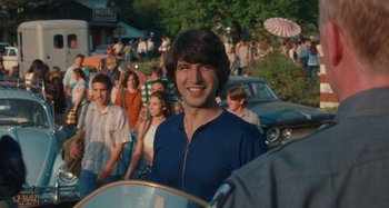 Movie still from “Taking Woodstock” (2009), directed by Ang Lee – A young man smiles while standing in front of a group of people; Medium shot, Over the shoulder angle